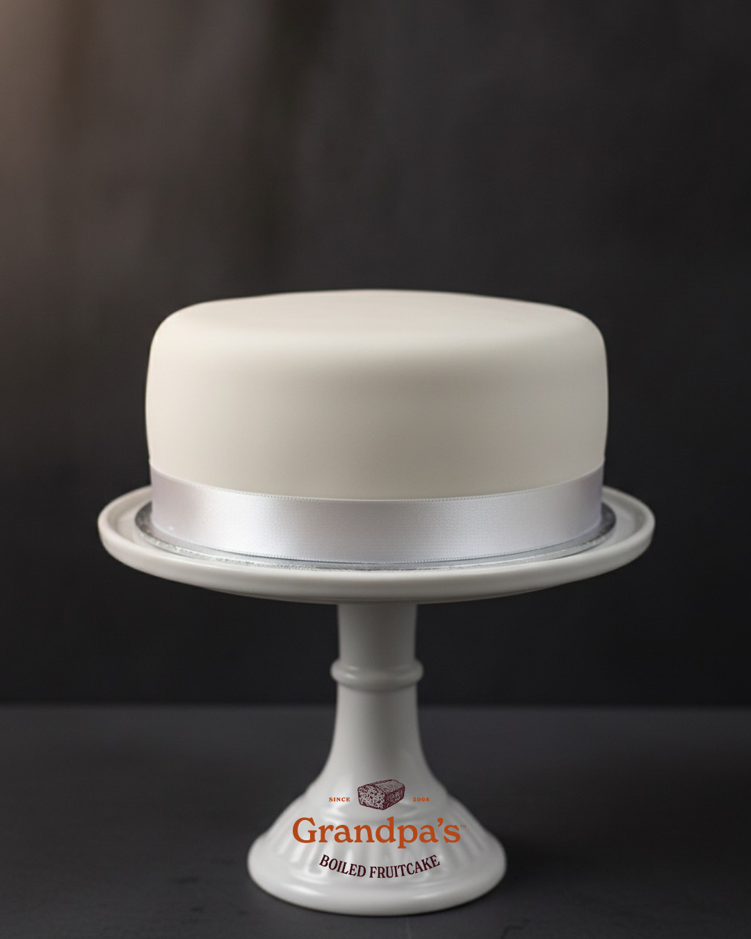 White fruit cake on a white cake stand with 'Grandpa's' branding against a dark background